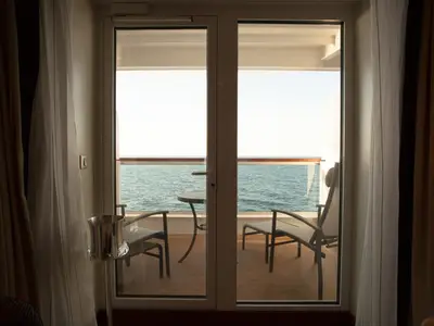Blick durch die Balkontür einer Kabine an Bord der Seabourn Encore auf den Balkon mit zwei Stühlen, einem Tisch und dem blauen Meer.