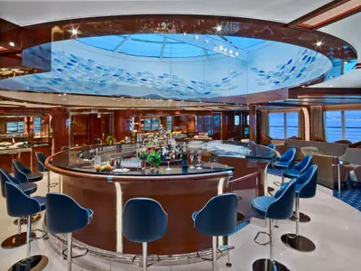 Die elegante Bar an Bord der Seabourn Encore verfügt über einen runden Tresen, blaue Barhocker und eine kunstvolle Fisch-Deckeninstallation.