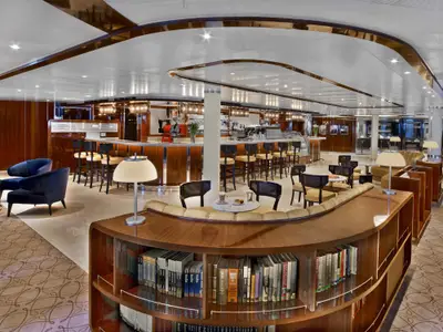 Ein stilvoller Lounge-Bereich an Bord der Seabourn Encore mit einem geschwungenen Bücherregal, gemütlichen Sesseln und einer Bar im Hintergrund.