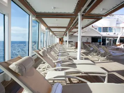 Reihe von eleganten Sonnenliegen an Bord der Seabourn Encore auf dem geschützten Deck mit Blick durch Glasscheiben auf das blaue Meer.