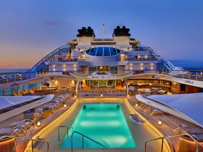 Der beleuchtete Poolbereich an Bord der Seabourn Encore in der Abenddämmerung mit Liegestühlen und Tischen.