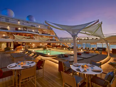 Blick über das beleuchtete Pooldeck an Bord der Seabourn Encore bei Sonnenuntergang mit Pool, Liegen und Tischen.