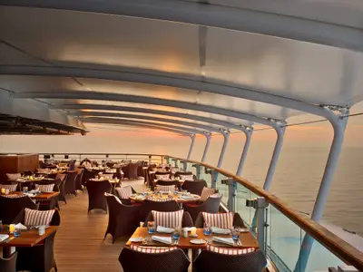 Das Restaurant The Colonnade an Bord der Seabourn Encore mit gedeckten Tischen im Außenbereich bei Sonnenuntergang über dem Meer.