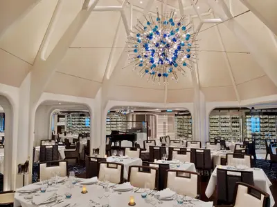 Das Hauptrestaurant an Bord der Seabourn Encore mit weiß gedeckten Tischen, edlen Stühlen und einem auffälligen blauen Glaskronleuchter.
