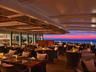 Eingedeckte Tische im Restaurant The Colonnade an Bord der Seabourn Encore bei einem malerischen Sonnenuntergang über dem Meer.