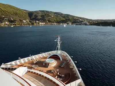 Blick von oben auf den Bug der Seabourn Encore mit einem Whirlpool und Liegestühlen auf dem Holzdeck vor einer grünen Küstenlandschaft.