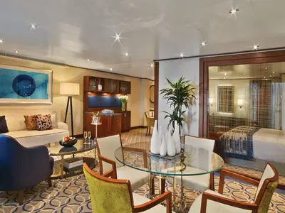 Elegante Owner's Suite an Bord der Seabourn Encore mit stilvollem Wohnbereich, rundem Glastisch und Blick durch eine Glasscheibe in das Schlafzimmer.