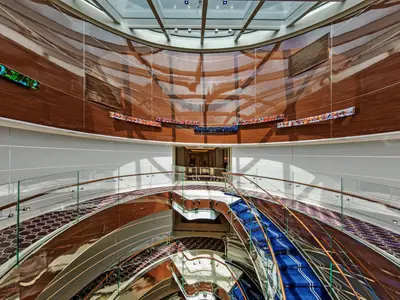 Blick in das Atrium an Bord der Seabourn Ovation mit geschwungenen Holzdecken, einer blauen Treppe und eleganten Glasgeländern.