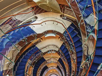 Blick von oben in das elegant geschwungene, spiralförmige Treppenhaus an Bord der Seabourn Ovation mit blauen Teppichstufen und Glasgeländer.