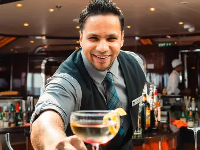 Nahaufnahme eines lächelnden Barkeepers an Bord der Seabourn Ovation, der dem Gast einen servierbereiten Cocktail reicht.