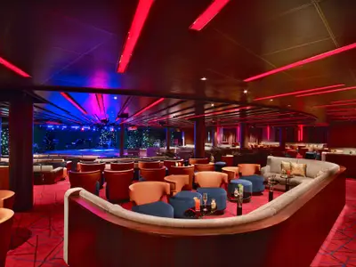 Elegante Club-Lounge an Bord der Seabourn Ovation mit gemütlichen Sesseln, kleinen Cocktailtischen, rotem Teppich und einer Veranstaltungsbühne.