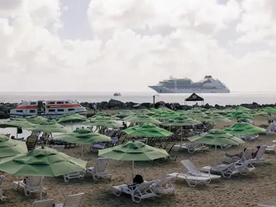Sandstrand mit vielen grünen Sonnenschirmen und Liegen, links ein Tenderboot der Seabourn Ovation und im Hintergrund das Hauptschiff.