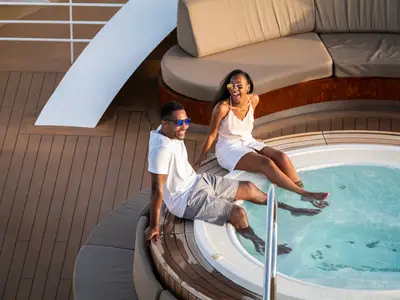 Ein lächelndes Paar sitzt am Rand eines Whirlpools auf dem Außendeck an Bord der Seabourn Ovation und lässt die Beine ins Wasser hängen.