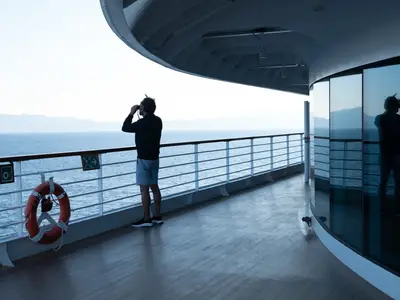 Ein Mann steht mit einem Fernglas an der Reling auf dem Außendeck an Bord der Seabourn Ovation und blickt auf das offene Meer.