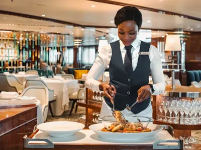 Eine lächelnde Servicemitarbeiterin in Weste serviert ein warmes Gericht im eleganten Restaurant an Bord der Seabourn Ovation.