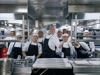 Gruppenfoto der Köche und Küchencrew in weißen Uniformen und schwarzen Schürzen in der Großküche an Bord der Seabourn Ovation.