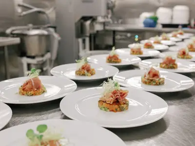 Reihen von weißen Tellern mit kunstvoll angerichtetem Schinken und Gemüse in der Küche an Bord der Seabourn Ovation.