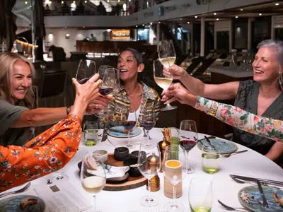 Eine Gruppe von Frauen stößt gut gelaunt mit Weingläsern an einem elegant gedeckten Tisch an Bord der Seabourn Ovation an.