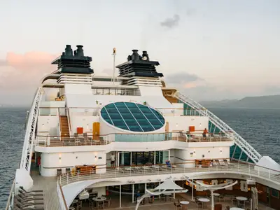 Blick auf das elegante Außendeck der Seabourn Ovation mit einem runden Glas-Oberlicht, Liegestühlen, Schornsteinen und dem Meer im Hintergrund.