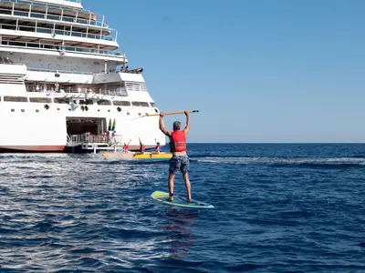 Ein Mann auf einem SUP-Board vor dem Heck der Seabourn Ovation, während im Hintergrund ein Bananenboot fährt.