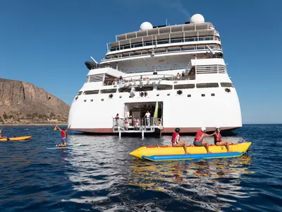 Gäste vergnügen sich beim Wassersport vor der Marina an Bord der Seabourn Ovation mit einem Bananenboot und SUP.