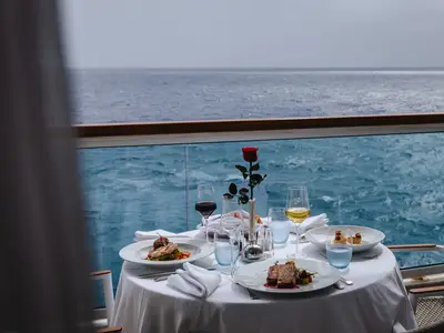 Ein gedeckter Tisch mit Speisen, Wein und einer roten Rose auf dem Balkon einer Suite an Bord der Seabourn Ovation vor Meereshintergrund.