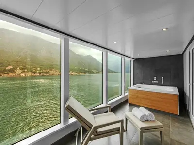 Luxuriöse Wellness-Oase in einer Suite an Bord der Seabourn Ovation mit Whirlpool, Liegestuhl und Panoramablick durch bodentiefe Fenster auf grüne Berge und Meer.