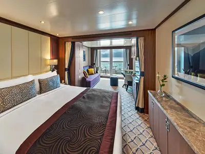 Blick durch eine elegante Suite an Bord der Seabourn Ovation mit großem Bett, Wohnbereich und Balkon mit Meerblick.