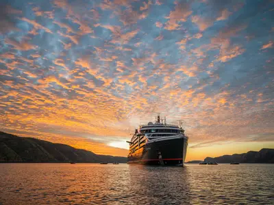 Das Expeditionsschiff Seabourn Pursuit liegt bei Sonnenuntergang vor einer Küste auf dem Wasser, umgeben von kleinen Zodiac-Booten.