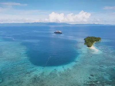 Luftaufnahme der Seabourn Pursuit in einer tiefblauen Lagune, umgeben von tropischen Inseln und Korallenriffen.