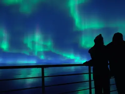Zwei Silhouetten an Bord der Seabourn Pursuit blicken auf die leuchtenden grünen Polarlichter am Nachthimmel über dem ruhigen Meer.