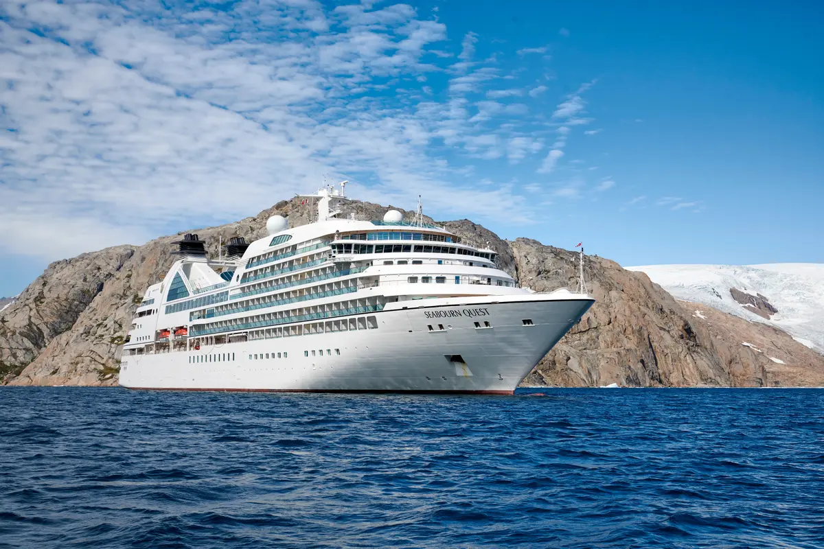 Die weiße Seabourn Quest liegt auf blauem Wasser vor felsigen Klippen und einem Gletscher unter blauem Himmel.