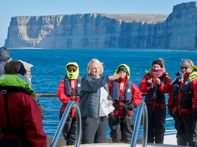 Eine Gruppe in roten Sicherheitsjacken lauscht dem Guide an Bord der Seabourn Quest vor einer riesigen Klippe im Meer.