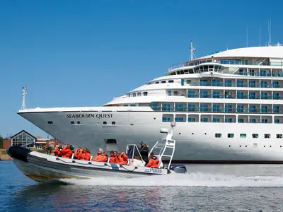 Ein Zodiac-Schnellboot mit Passagieren in orangefarbenen Jacken fährt vor dem großen Kreuzfahrtschiff Seabourn Quest im blauen Wasser.