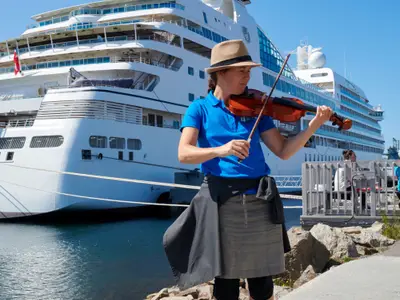 Eine Musikerin mit Hut spielt Violine auf dem Pier vor dem luxuriösen Kreuzfahrtschiff Seabourn Quest.