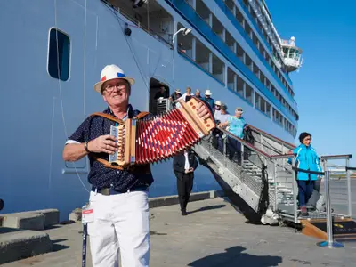 Ein Musiker mit Hut spielt Akkordeon an der Gangway des Luxusschiffs Seabourn Quest, während Passagiere im Hafen von Bord gehen.