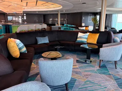 Eine moderne Lounge an Bord der Seabourn Quest mit einem großen, dunklen Sofa, farbigen Kissen, runden Glastischen und einem gemusterten Teppich.