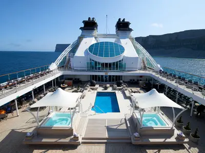 Symmetrische Aufnahme des eleganten Pooldecks an Bord der Seabourn Quest mit zwei Whirlpools, einem Pool und Klippen im Hintergrund.