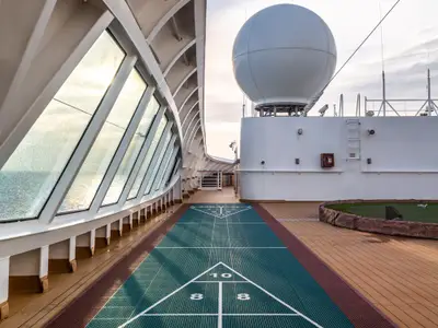 Ein grünes Shuffleboard-Spielfeld auf dem Holzdeck an Bord der Seabourn Quest, daneben eine große Radarkuppel und schräge Glasfenster.