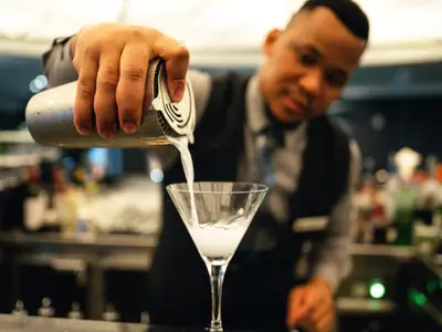 Nahaufnahme eines Barkeepers an Bord der Seabourn Sojourn, der einen weißen Cocktail aus einem Shaker in ein Martiniglas einschenkt.