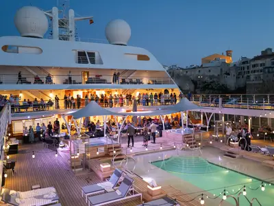 Gäste versammeln sich bei Abenddämmerung am beleuchteten Pooldeck an Bord der Seabourn Sojourn vor einer Altstadtkulisse.
