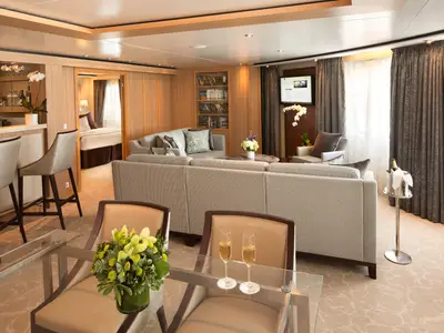 Einblick in eine elegante Suite an Bord der Seabourn Sojourn mit grauem Sofa, Esstisch mit Sektgläsern und separatem Schlafbereich.