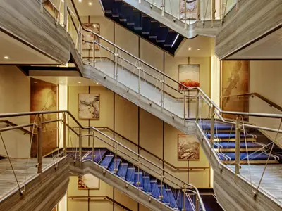 Das moderne, mehrstöckige Treppenhaus an Bord der Seabourn Venture mit blauem Teppich, Holzverkleidungen und Wandbildern.