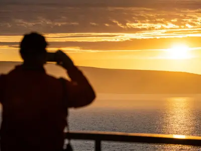 Silhouette eines Gastes, der den goldenen Sonnenuntergang über dem Meer an Bord der Seabourn Venture fotografiert.
