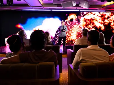 Gäste sitzen im Discovery Center an Bord der Seabourn Venture und lauschen einem Vortrag vor einer großen, bunten LED-Leinwand.