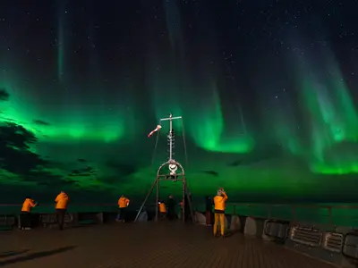 Passagiere in orangefarbenen Jacken beobachten spektakuläre grüne Polarlichter am Nachthimmel an Bord der Seabourn Venture.