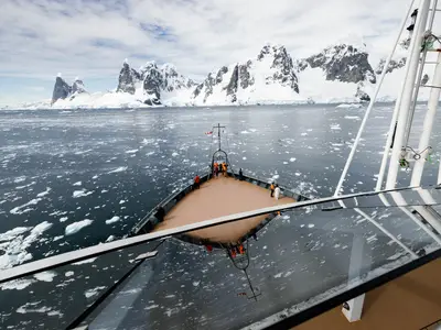 Blick vom Aussichtsdeck der Seabourn Venture auf den Bug des Schiffes, das durch Treibeis vor schneebedeckten Bergen in der Antarktis fährt.