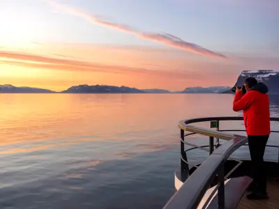 Ein Passagier in roter Jacke fotografiert an Bord der Seabourn Venture einen malerischen Sonnenuntergang über dem Polarmeer und fernen Bergen.