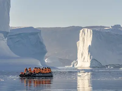 Passagiere in orangefarbenen Parkas fahren mit einem Zodiac der Seabourn Venture an riesigen Eisbergen in Grönland vorbei.
