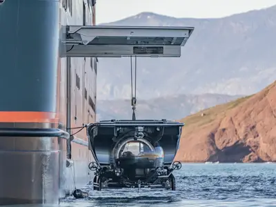 Ein U-Boot wird von einer Kranvorrichtung an Bord der Seabourn Venture ins Meer gelassen, im Hintergrund rötliche Berge.